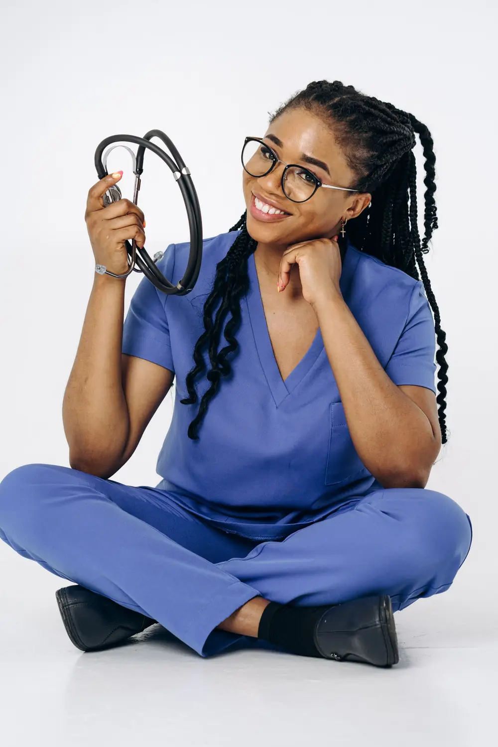 A medical student in scrubs sitting on the floor, smiling and holding a stethoscope.